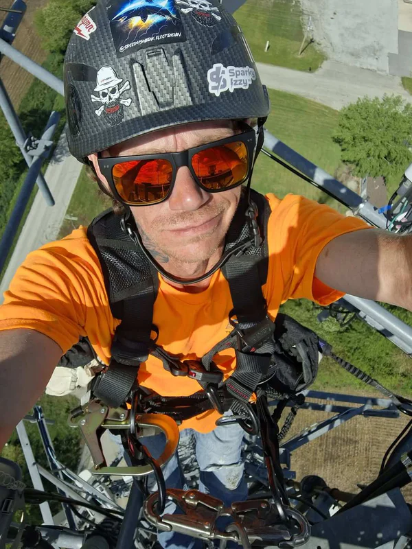 Telecom tower climber in full fall protection gear and helmet taking a selfie from high altitude on a cell tower. Wearing hi-vis orange shirt and safety harness with carabiners, overlooking ground below. Industrial tower work and height safety PPE.